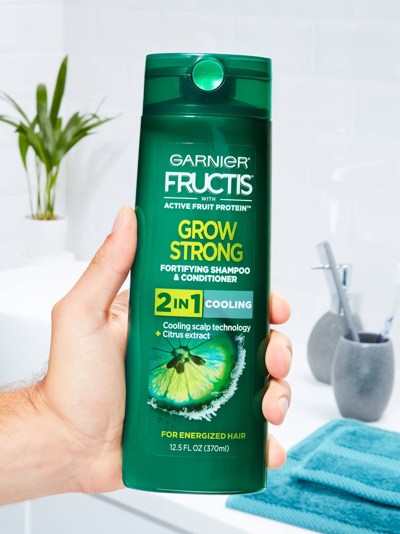 Hand holding Cooling 2-In-1 Shampoo and Conditioner for Men in a white bathroom with a blue towel, silver cups with toothbrushes, and a plant.
